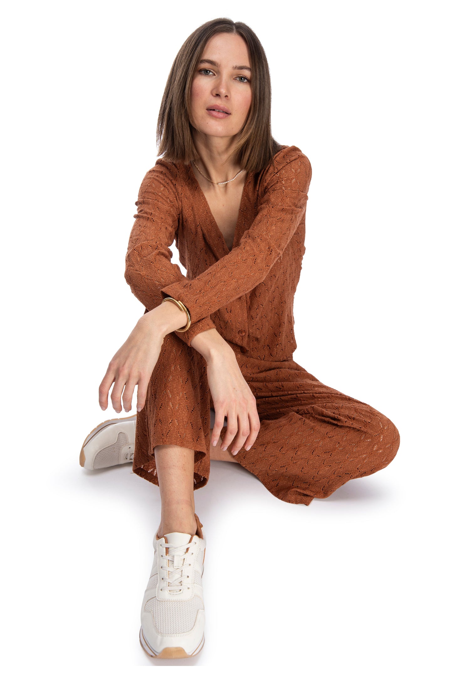 A woman in a textured brown dress with a relaxed pose sits against a white background, exuding a casual, confident style complemented by trendy white sneakers and her B Collection by Bobeau LS Crop Bttn Cardigan.