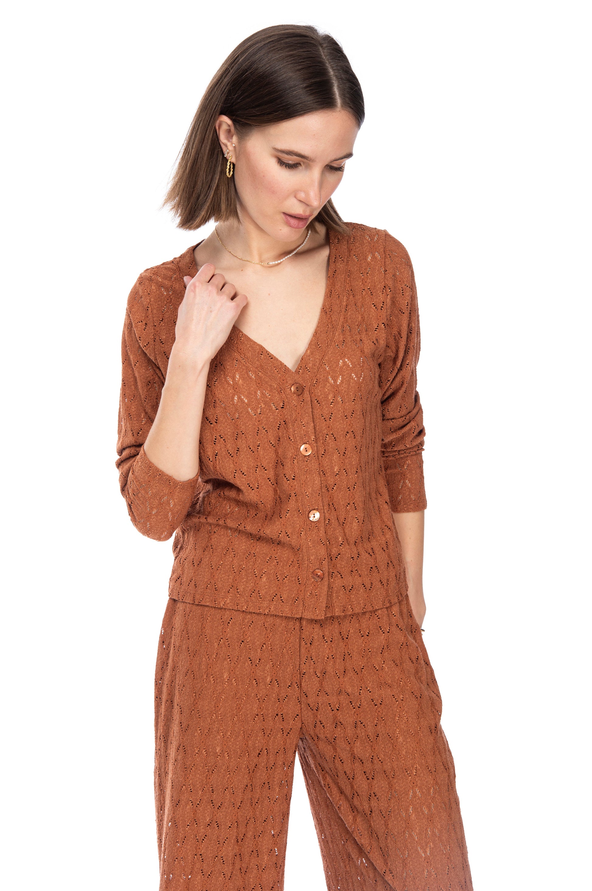 A woman in a stylish, textured terracotta LS Crop Bttn Cardigan and Gilly ankle-length pants looking down and to the side, lightly adjusting her collar.