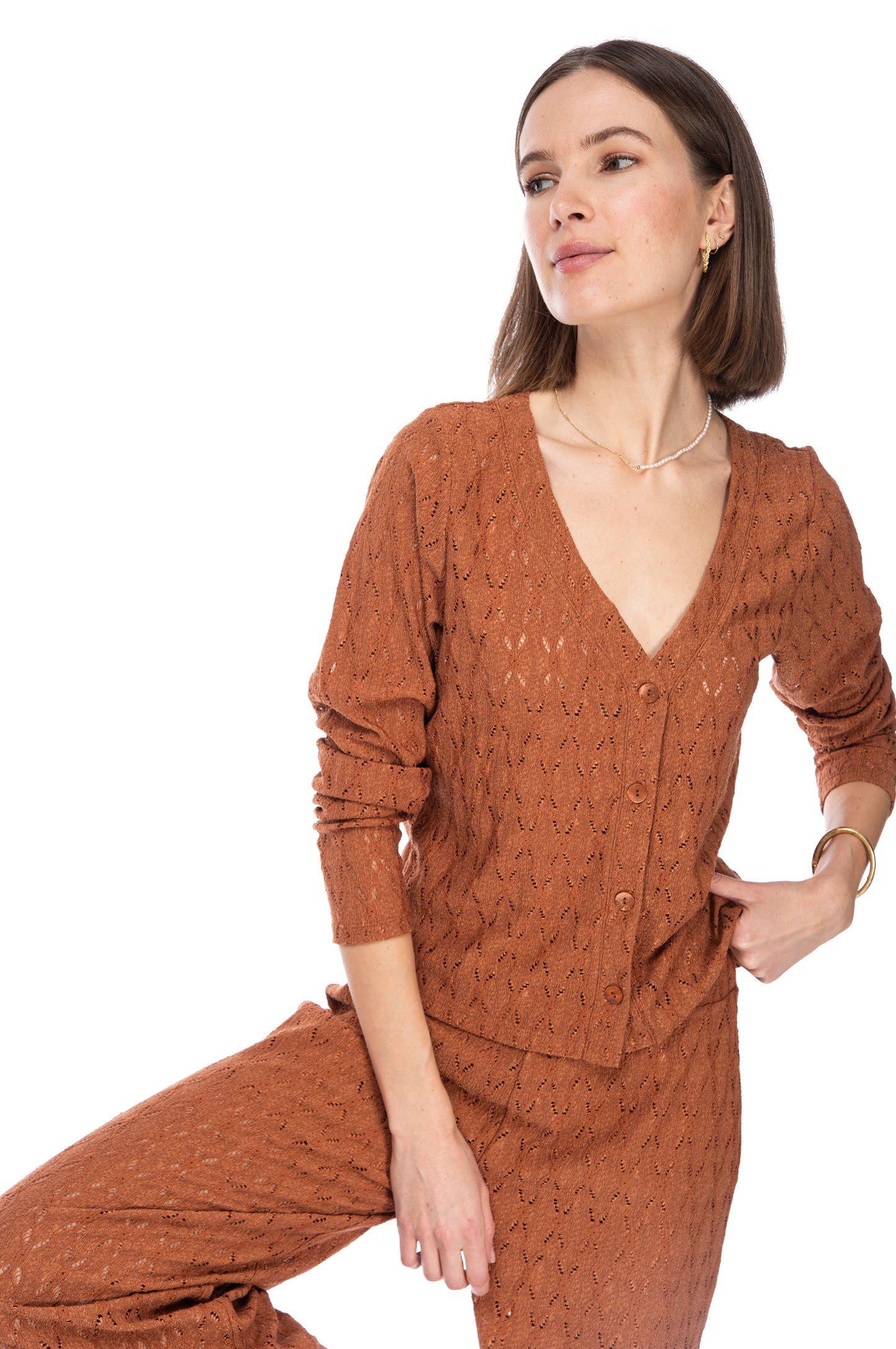 A woman posing confidently in a stylish rust-colored B Collection by Bobeau LS CROP BTTN CARDIGAN and matching Gilly ankle length pants, accessorized with a delicate necklace.