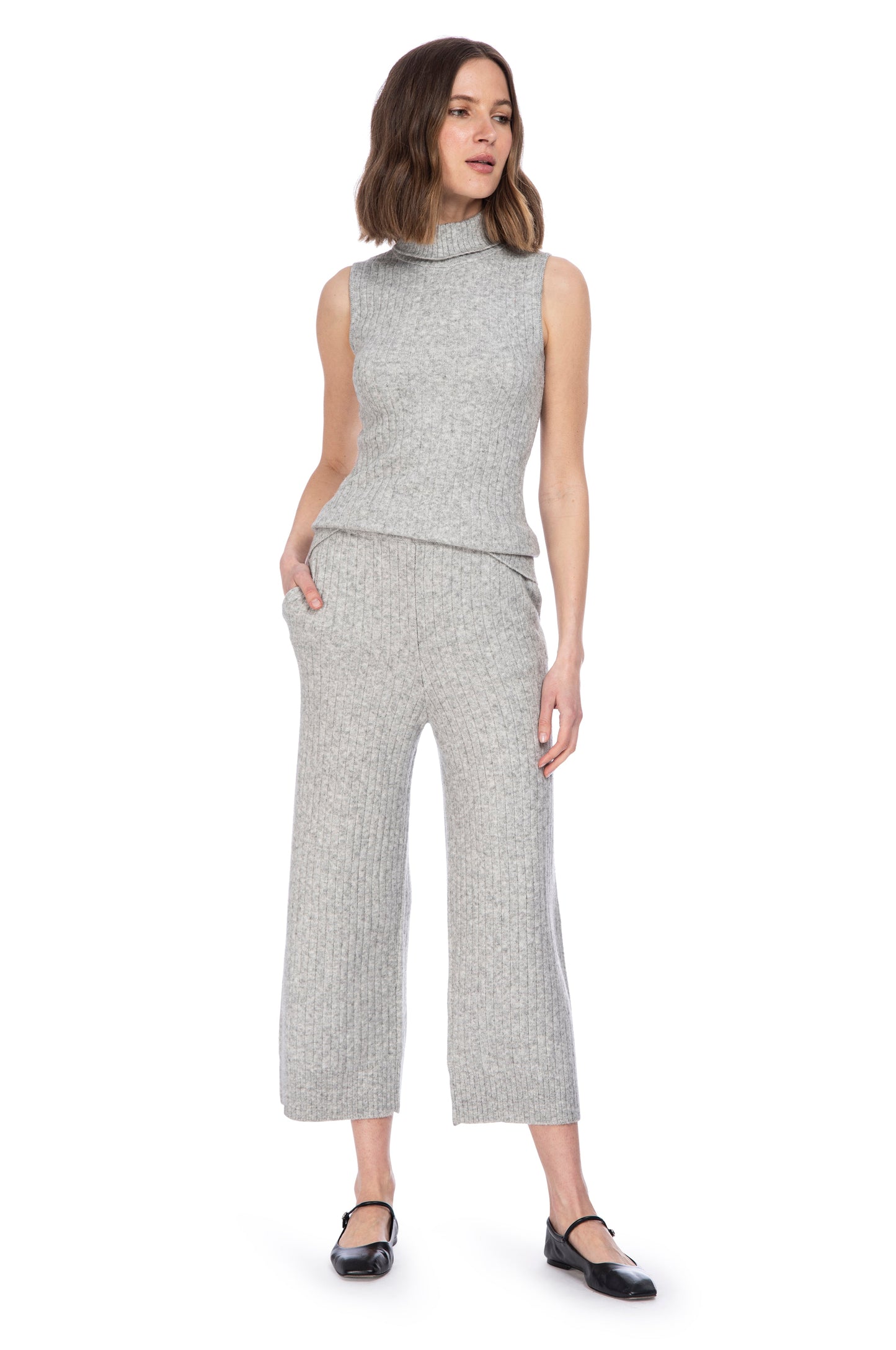 A woman posing in a casual grey "TURTLENECK SLVLSS NYLON BLEND" sleeveless turtleneck sweater by B Collection by Bobeau and matching cropped pants, paired with black shoes, exhibiting a modern and minimalist fashion style.