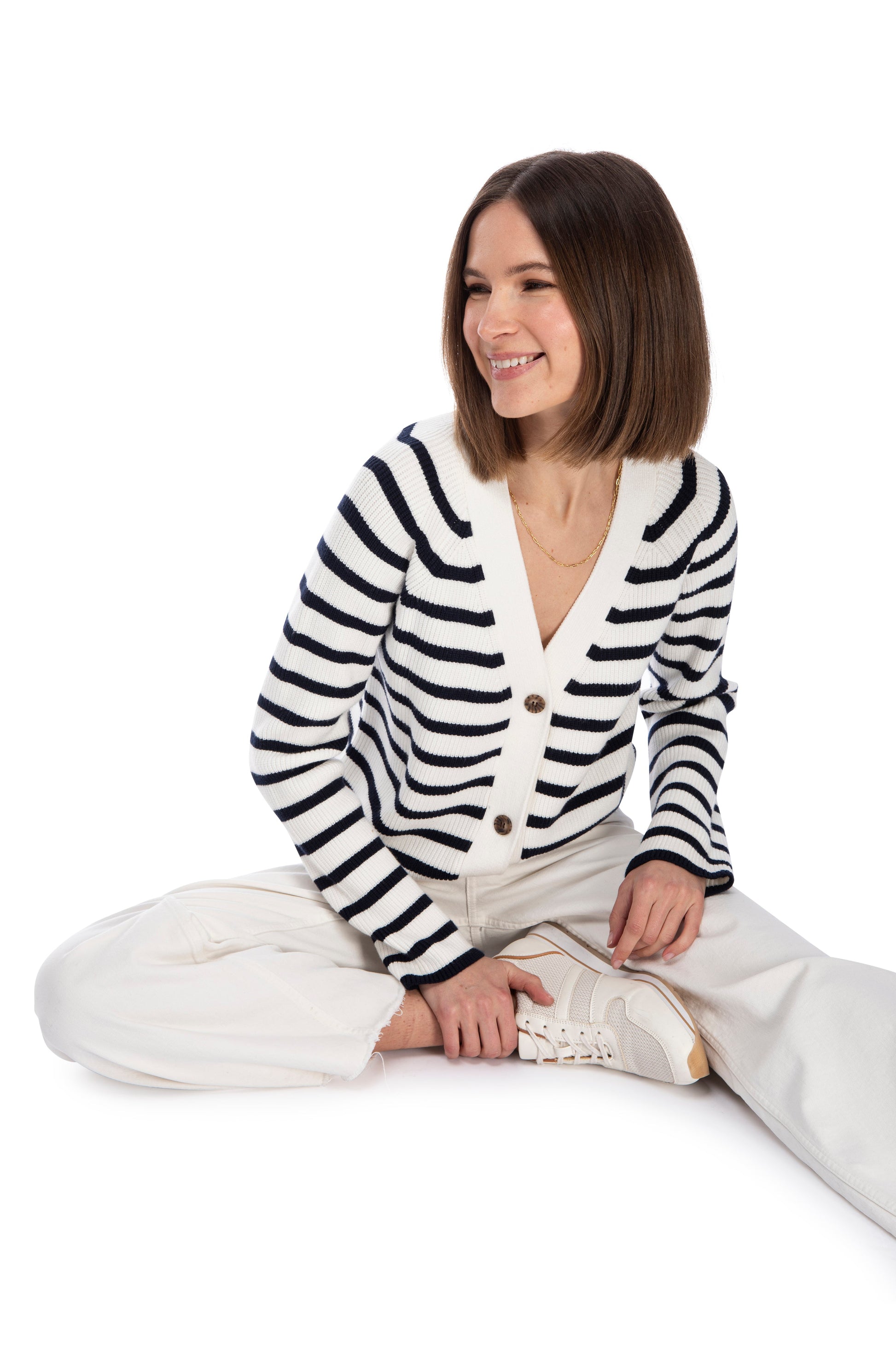 A woman with a joyful expression sitting cross-legged, wearing a B Collection by Bobeau LS STRIPE CARDIGAN made from super soft yarn, white pants, and sneakers, isolated on a white background.