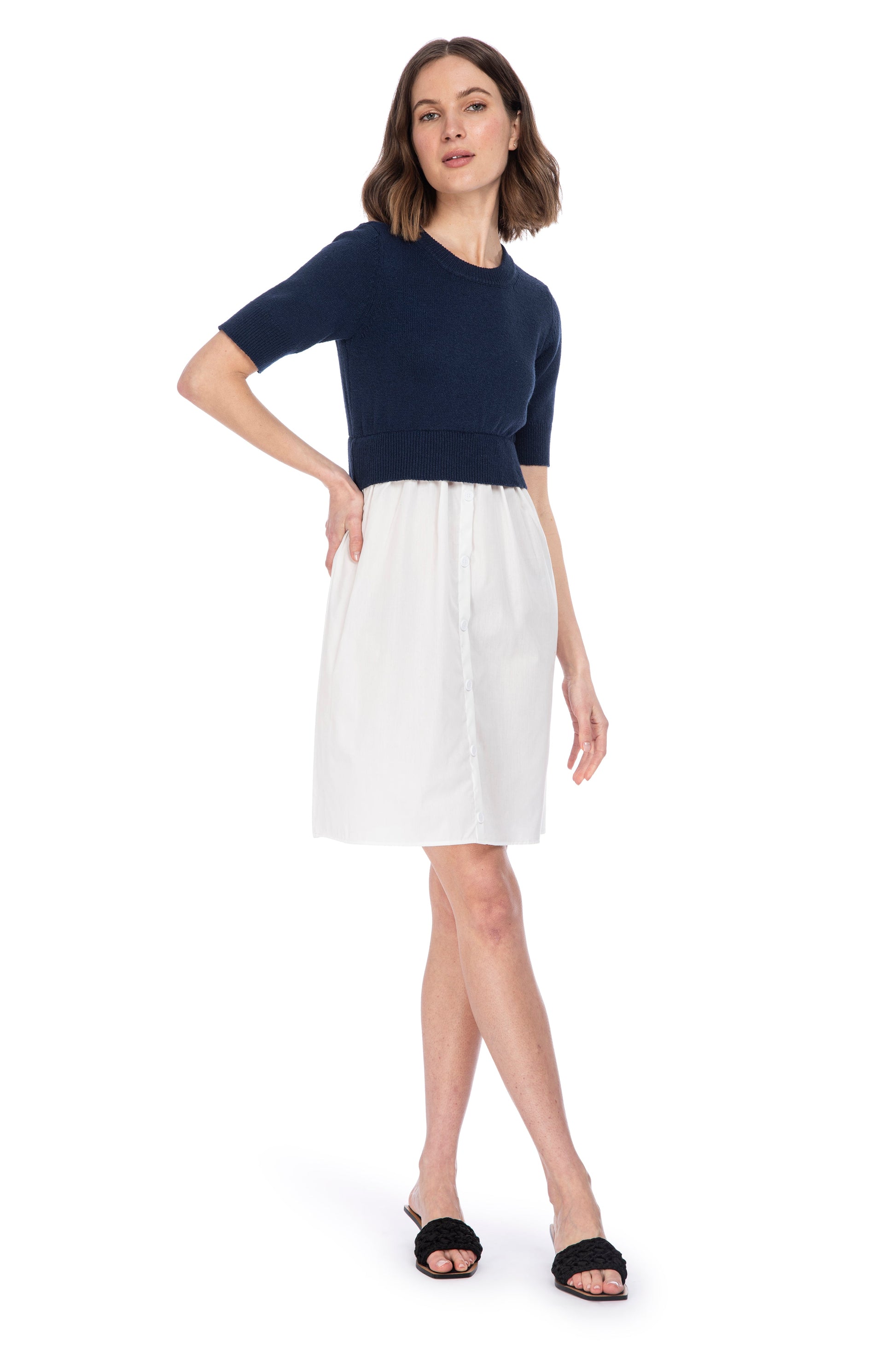 A woman modeling a casual SS Mixed Media Dress by B Collection by Bobeau with a navy blue top and a white poplin skirt, paired with simple black sandals.