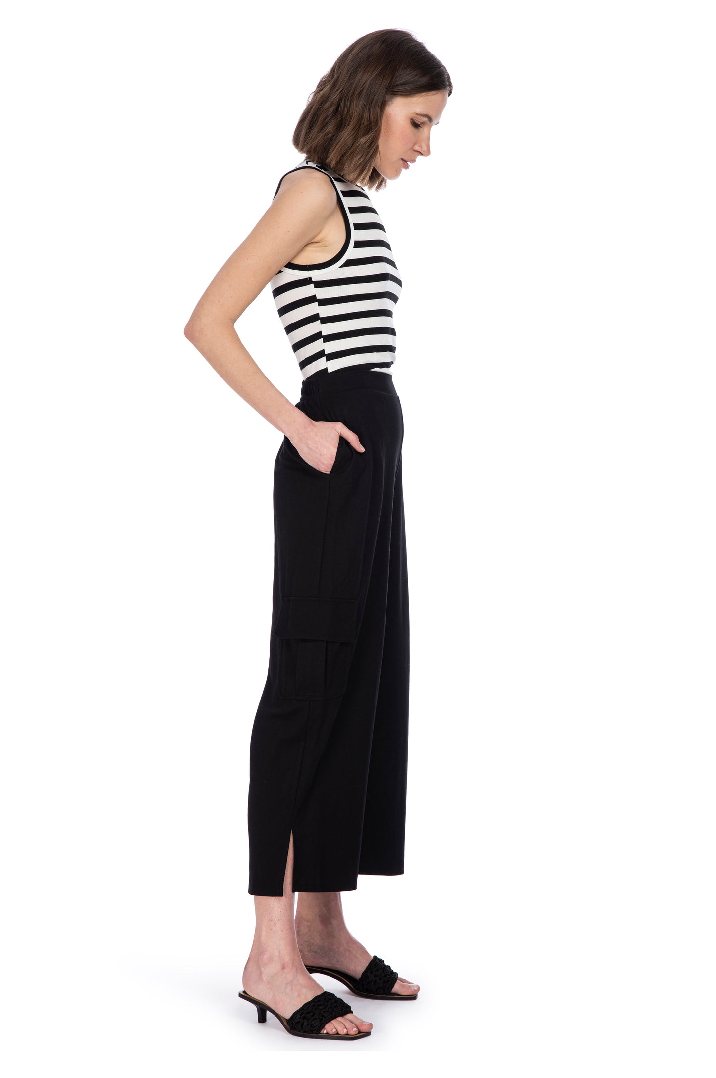 A woman in a chic B Collection by Bobeau black and white striped HIGH NECK TANK and high-waisted black trousers standing sideways, looking downwards with one hand on her hip.