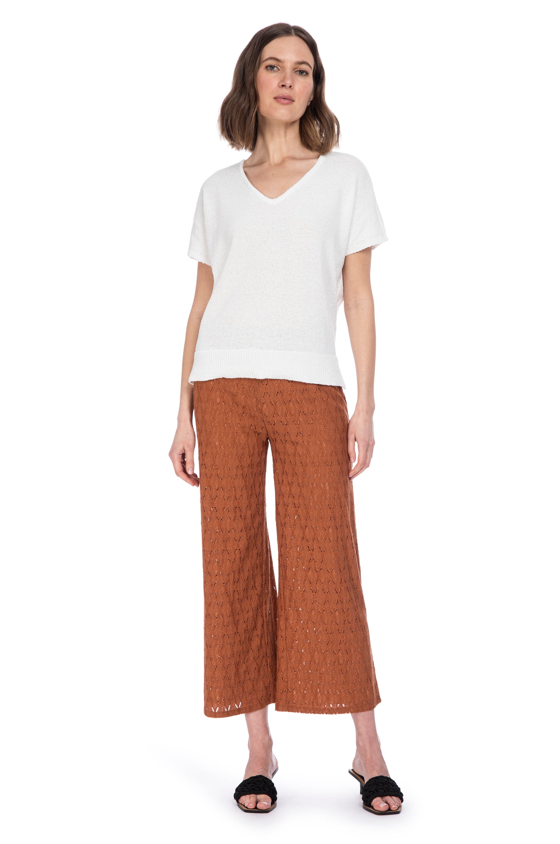 A woman stands confidently on a plain background, showcasing her fashion-forward outfit that includes white v-neck blouse paired with stylish brown lace-patterned culottes from B Collection by Bobeau, complemented by simple black open-toed sandals.