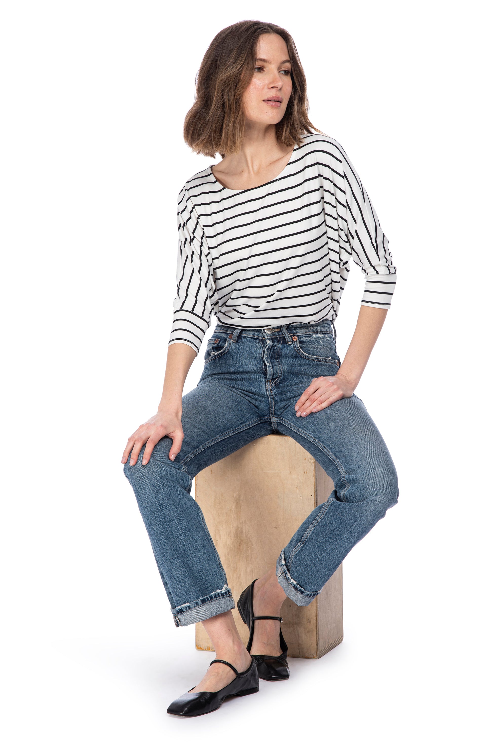Woman in a relaxed fit, B Collection by Bobeau 3/4 SLV DOLMAN BUTTER TOP and jeans seated on a wooden stool, looking away thoughtfully.