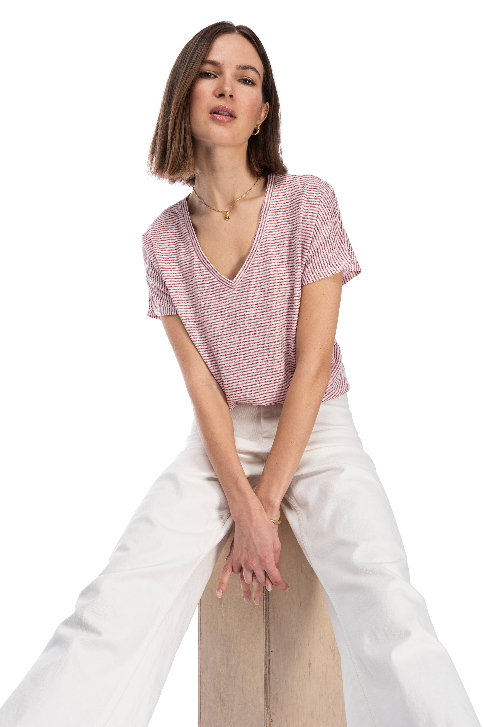Confident woman in a stylish CATY RELAXED TEE by B Collection by Bobeau and white pants posing casually while seated on a wooden stool against a white background.