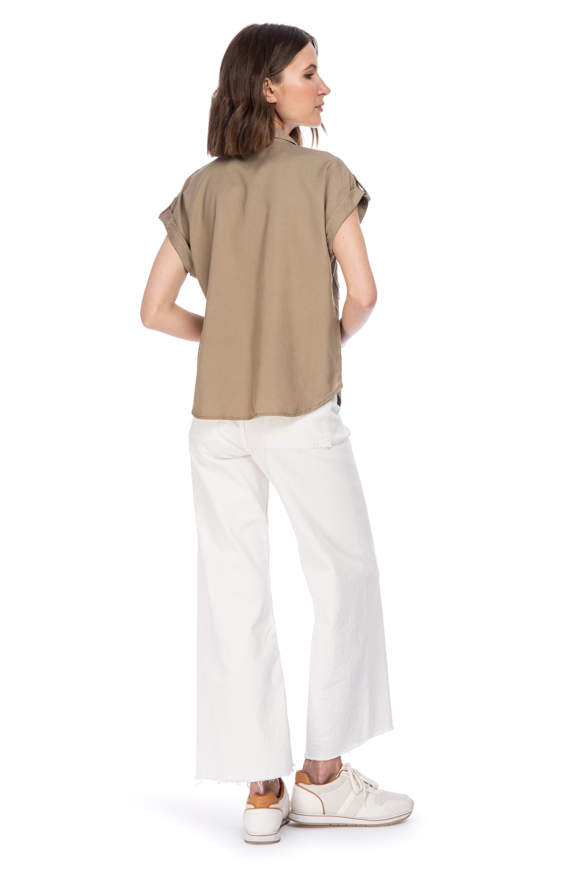 A woman viewed from the back, wearing a casual UTILITY BUTTON UP beige t-shirt from B Collection by Bobeau and white wide-leg pants, paired with white sneakers, looking to her left.