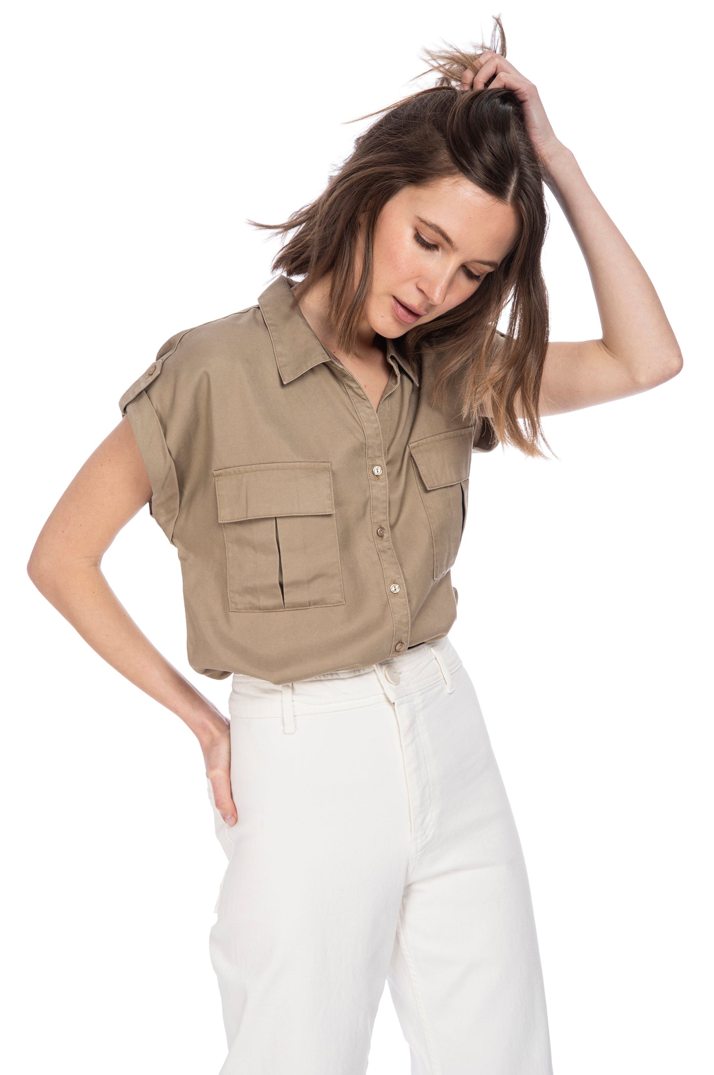A contemplative young woman in a casual B Collection by Bobeau Tencel khaki utility button-up top and white pants, standing with one hand on her hip and the other running through her hair.