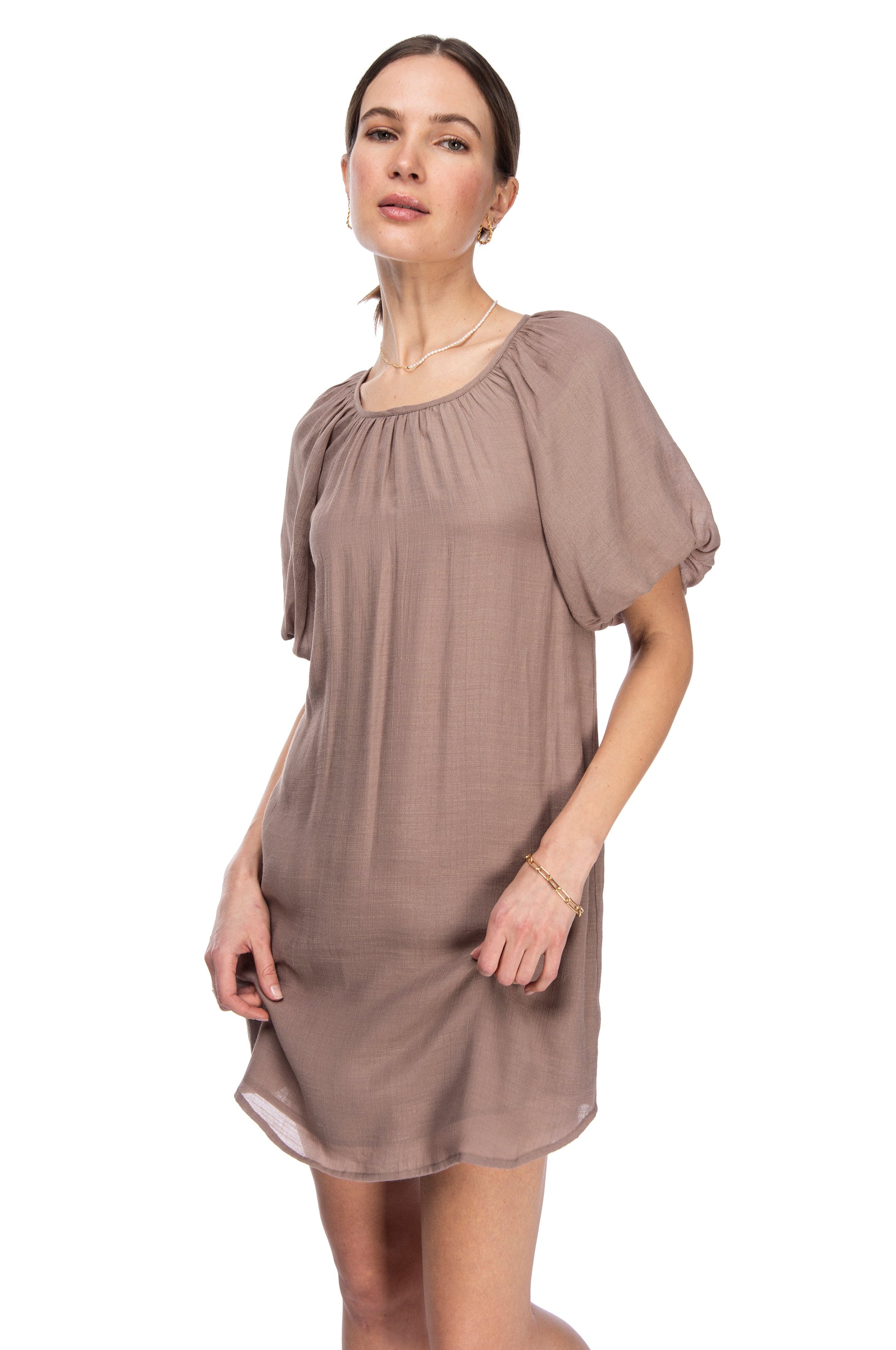 A woman standing against a white background, wearing a chic, short-sleeved, taupe gauze dress from B Collection by Bobeau with simple jewelry, looking directly at the camera with a neutral expression.
