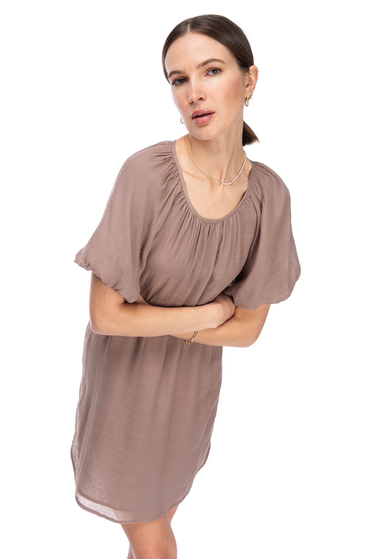 Woman standing against a white background wearing a loose-fitting taupe gauze dress with B Collection by Bobeau bubble hem sleeves, giving a curious look to the side.