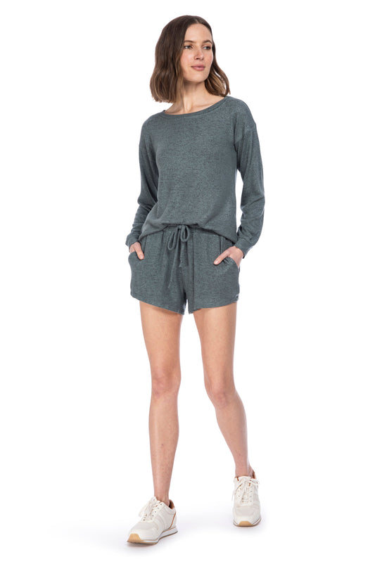 A woman stands confidently, modeling a casual gray long-sleeve B Collection by Bobeau COZY PULLOVER AND SHORT SET, paired with white sneakers.