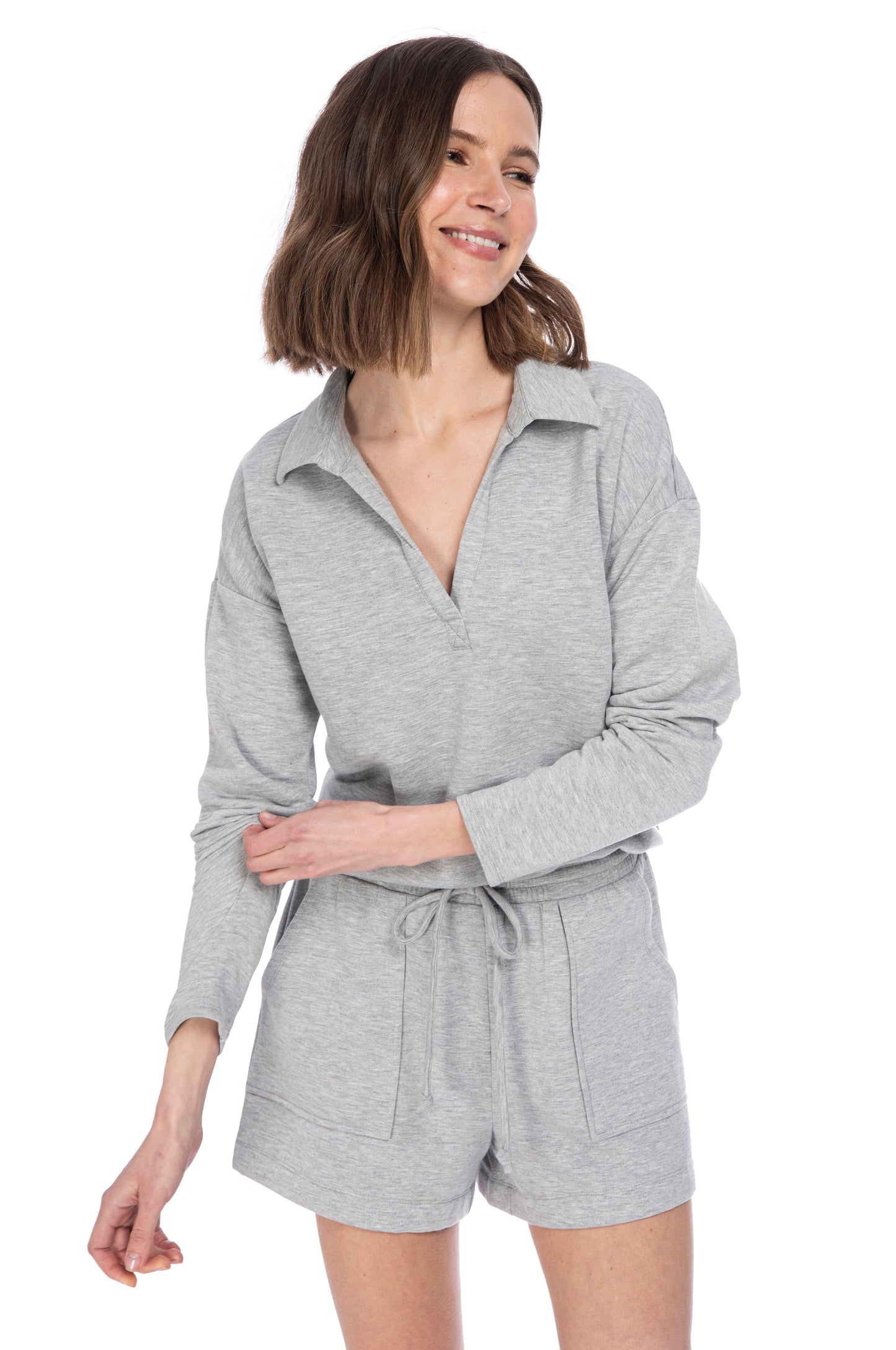 A woman in a casual gray B Collection by Bobeau FLEECE COLLAR TOP & SHORT SET, smiling subtly and posing against a white background.