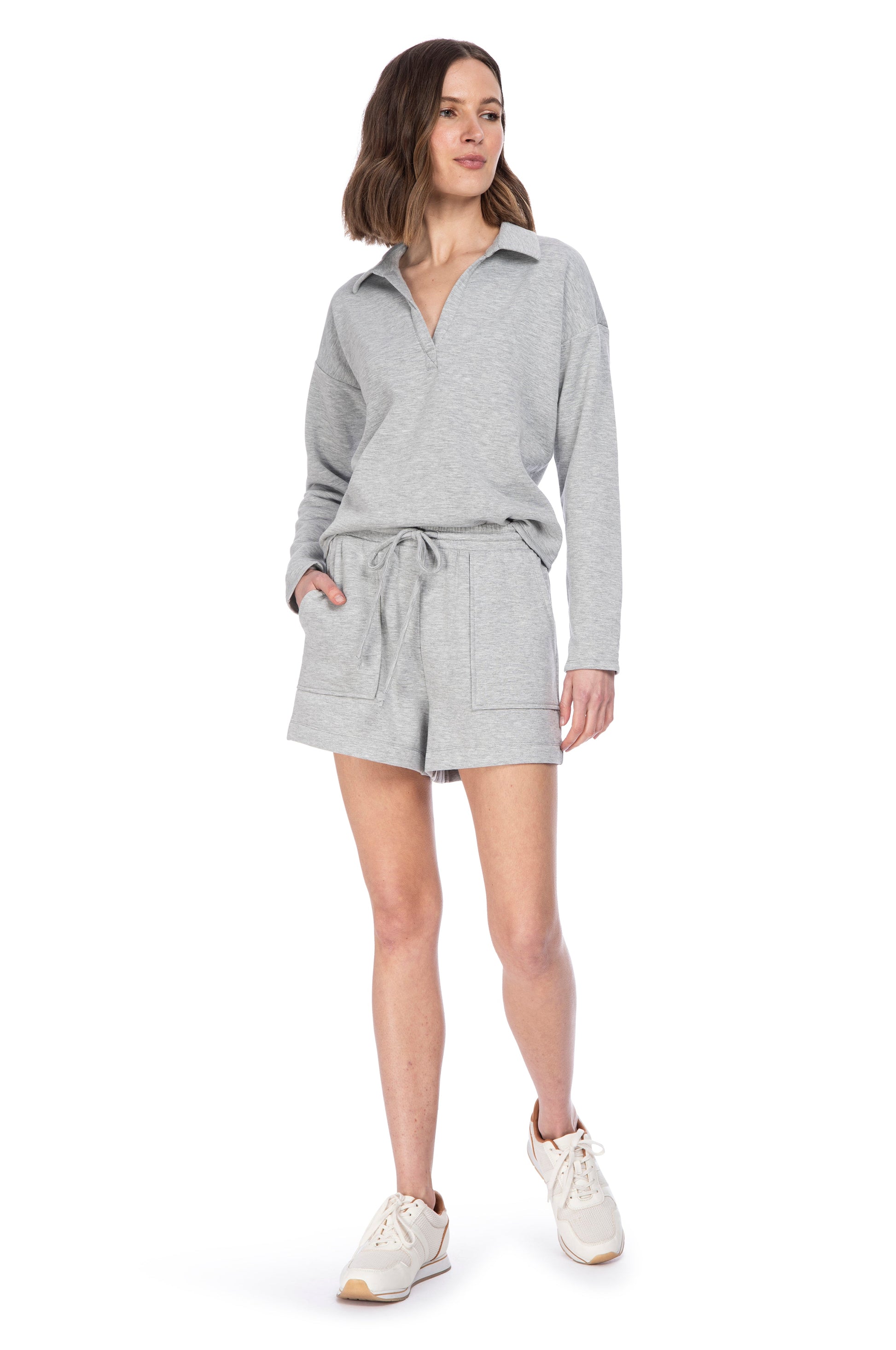 A woman in a casual grey B Collection by Bobeau Stella Fleece hooded playsuit with shorts and a drawstring waist, paired with white sneakers, stands confidently against a white background.