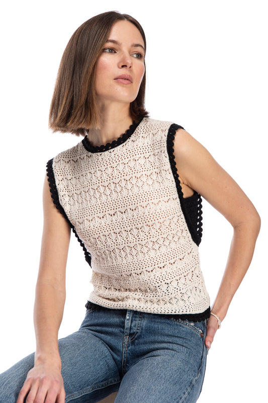 A young woman in a casual pose, wearing a sleeveless, beige, crochet knit sweater vest with scalloped edges and black trim from B Collection by Bobeau and classic blue jeans. She gives off a relaxed yet stylish vibe.