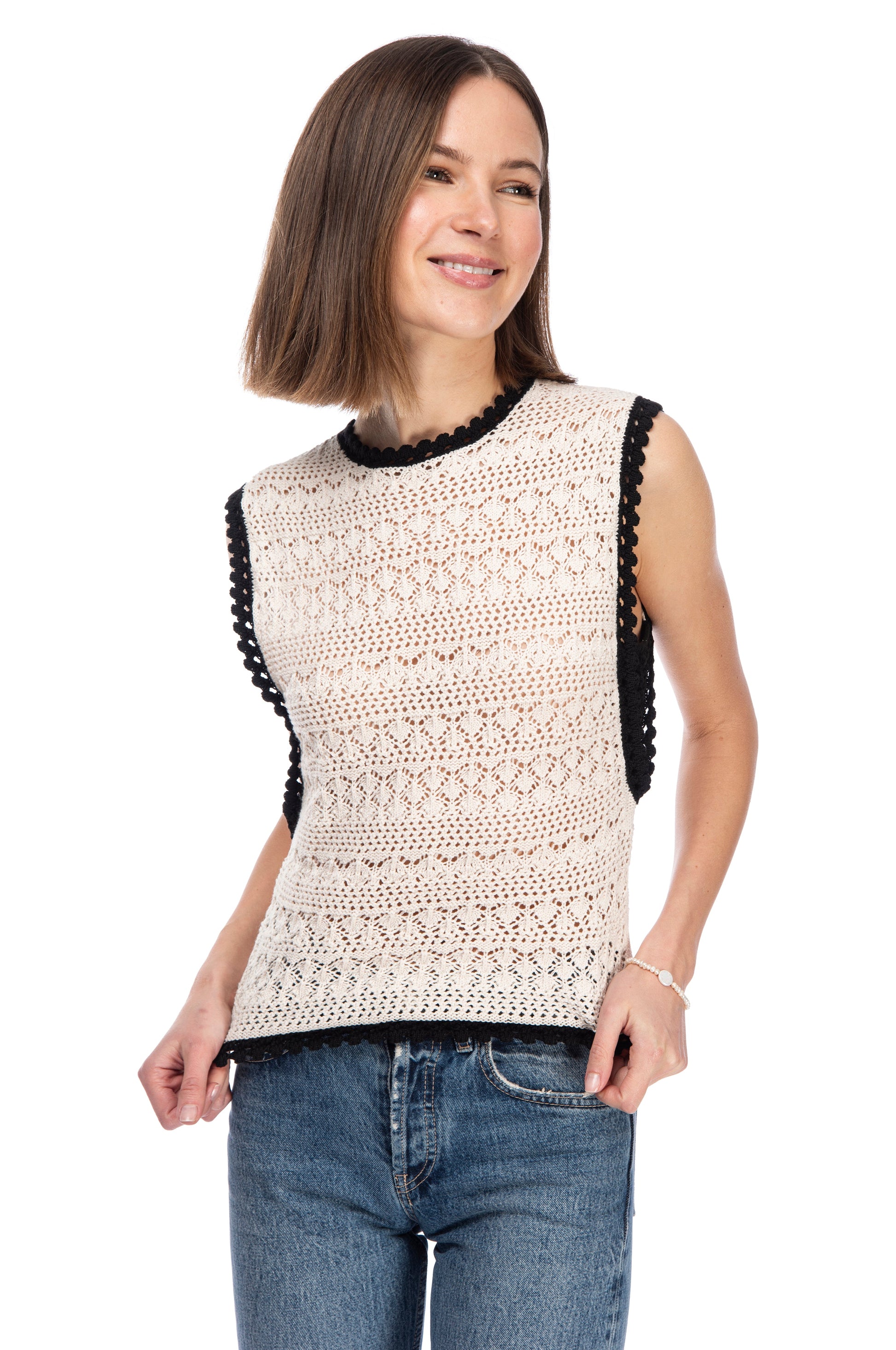 A woman in a beige SLVLSS CROCHET SWEATER TOP by B Collection by Bobeau with black trim and blue jeans standing against a white background, smiling gently.