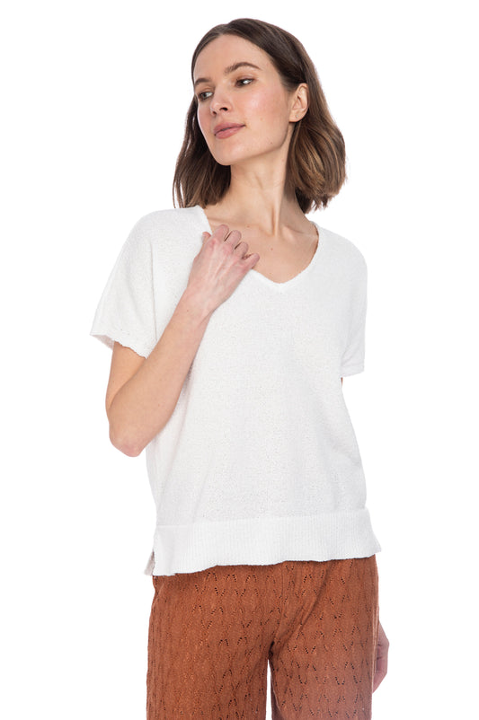 A woman in a casual B Collection by Bobeau V Neck Dolman Sweater and textured brown trousers, posing with one hand gently touching her chest.