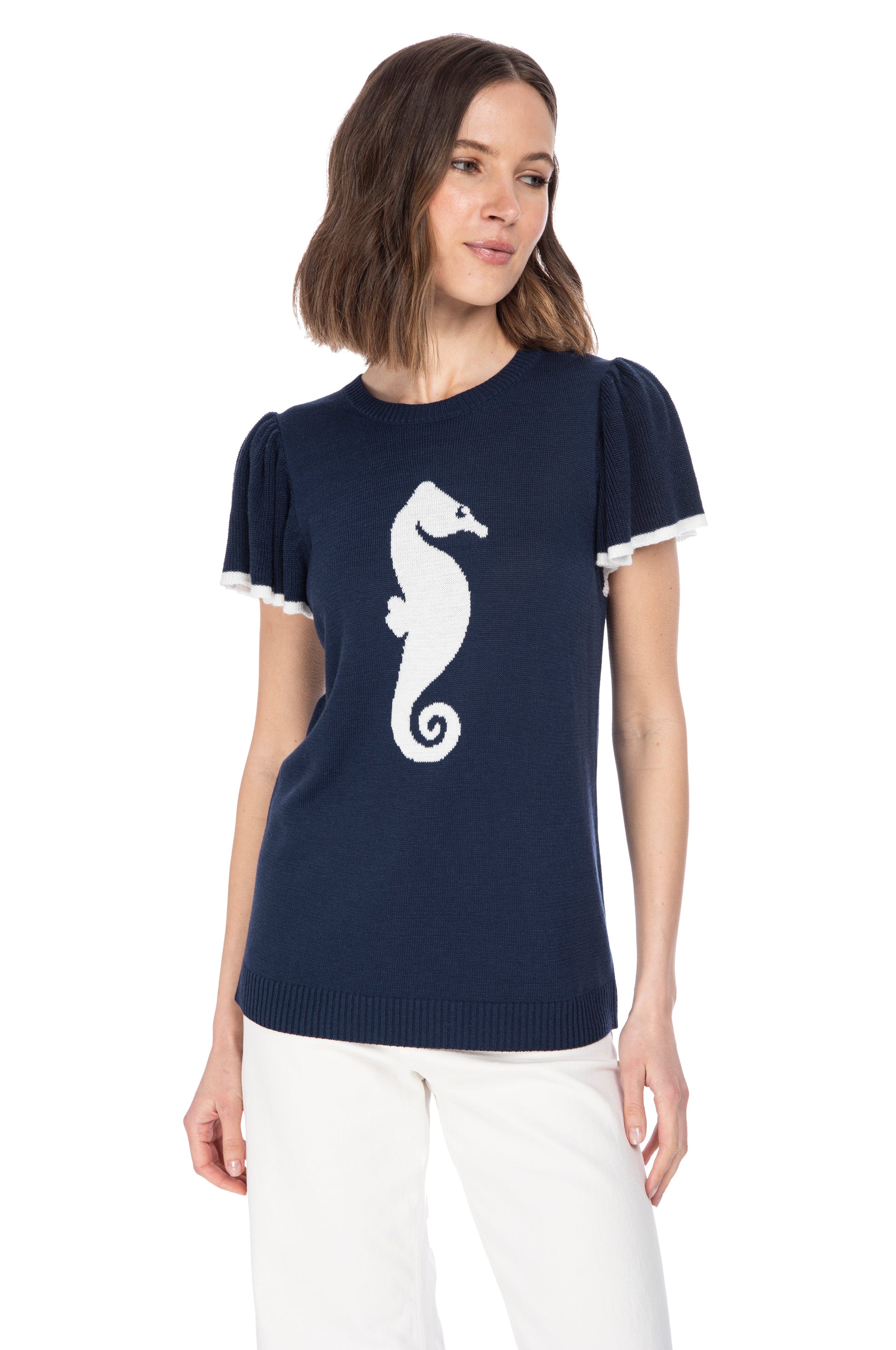 A woman in a chic navy blue SS Flutter Sleeve Top from B Collection by Bobeau with a white seahorse icon and stylish fringed sleeves, paired with crisp white pants, posing against a white background for a fresh, nautical look.