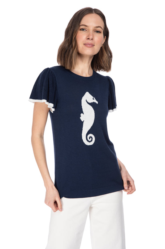 Woman in a chic navy blue SS flutter sleeve top with a white seahorse icon, paired with casual white trousers by B Collection by Bobeau.