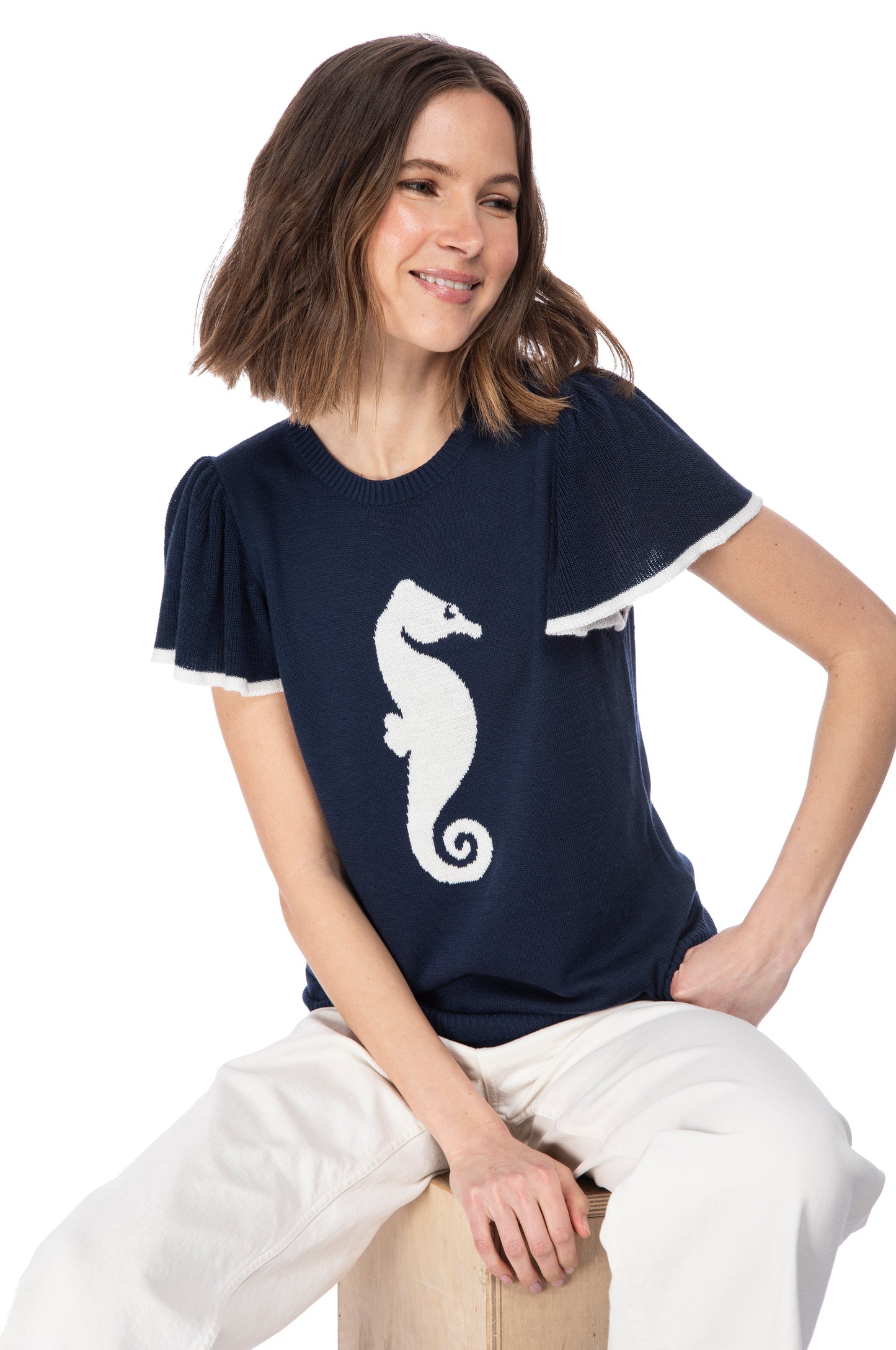 Woman in a navy blue SS flutter sleeve top by B Collection by Bobeau with a white seahorse icon design, smiling gently as she poses casually.