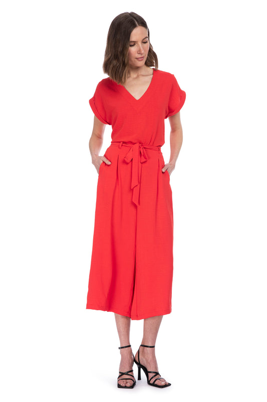 A woman posing in a stylish red jumpsuit with a TIE WAIST CROP TROUSER by B Collection by Bobeau, paired with elegant black heels, against a white background.