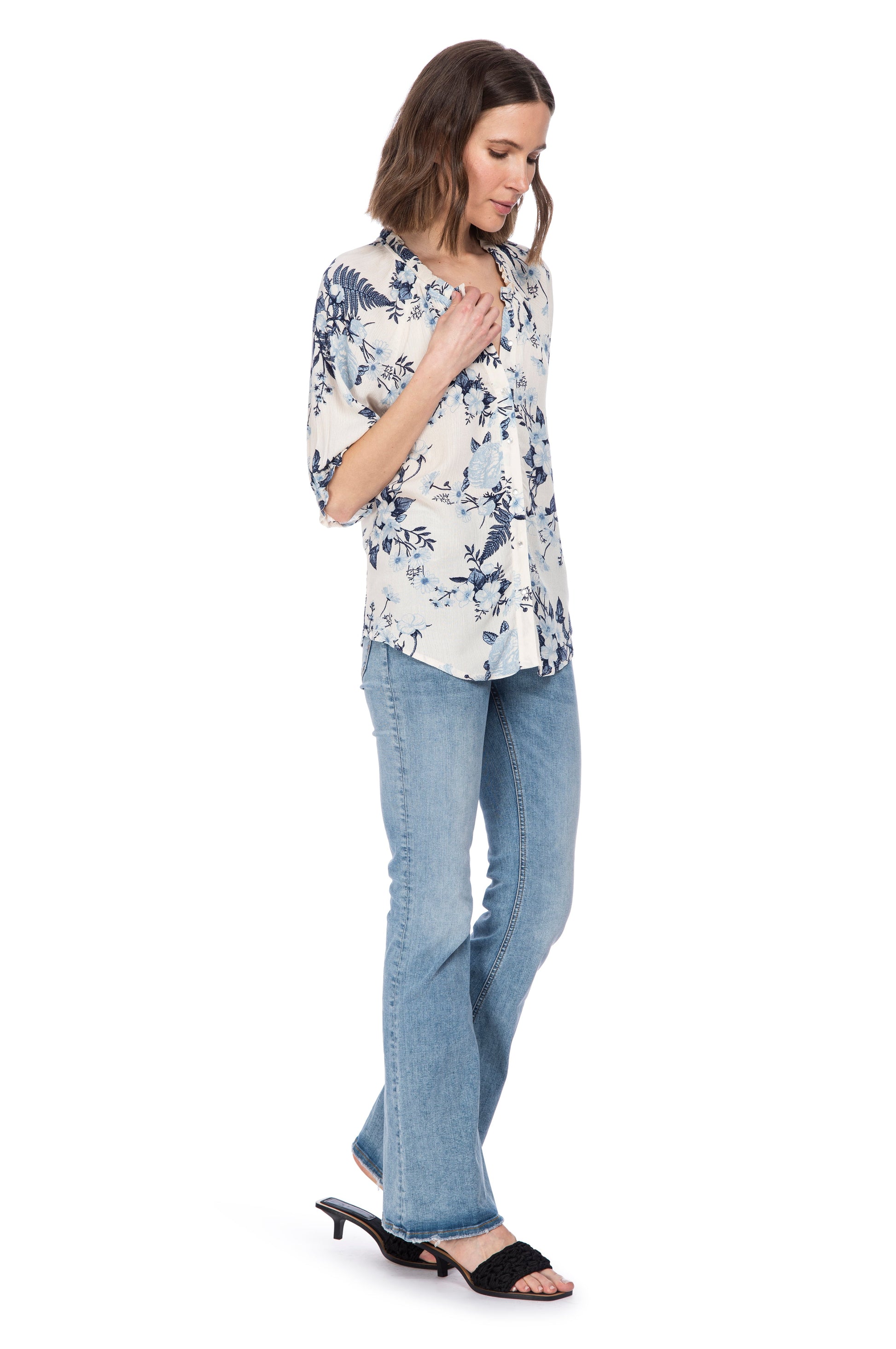 A woman in a BUTTON DOWN BLOUSE WITH RUFFLE NECK from B Collection by Bobeau with elasticated sleeves and jeans standing to the side while looking down thoughtfully.