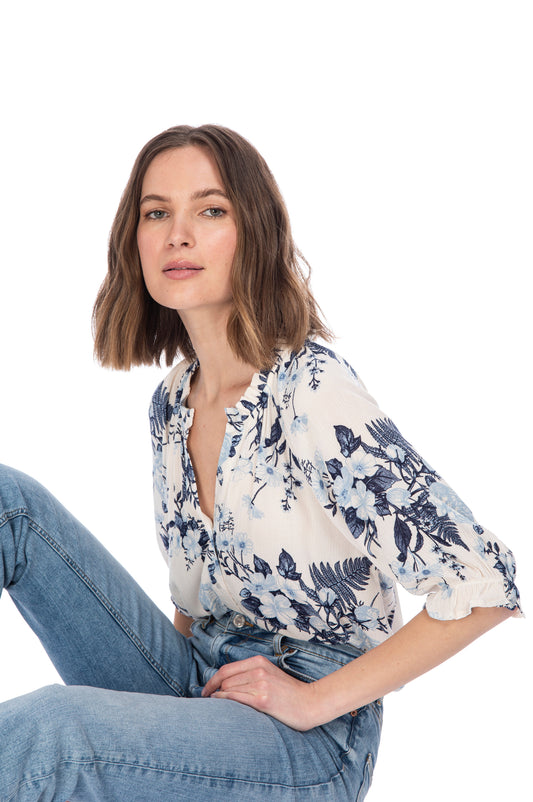 A woman in a BUTTON DOWN BLOUSE WITH RUFFLE NECK from B Collection by Bobeau with elasticated sleeves and jeans seated casually against a white background.