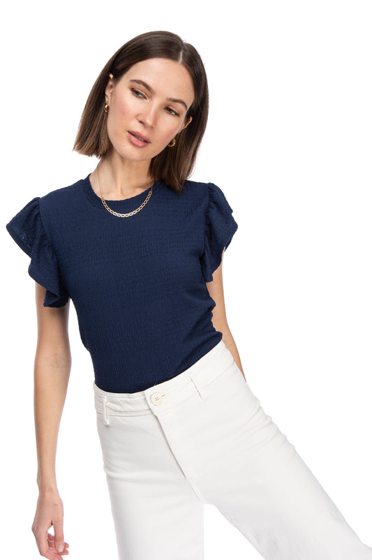 A woman with shoulder-length hair wearing a B Collection by Bobeau Bryce striped knit, RUFFLE SLV TOP, and white pants against a white background.