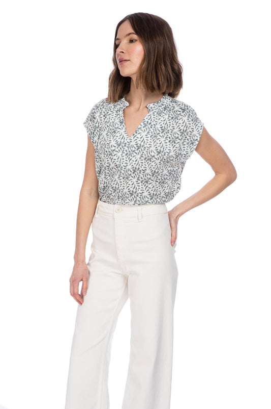 A woman in a B Collection by Bobeau CATY RUFFLE NECK TOP and white trousers stands with one hand on her hip, looking confidently to the side against a white background.