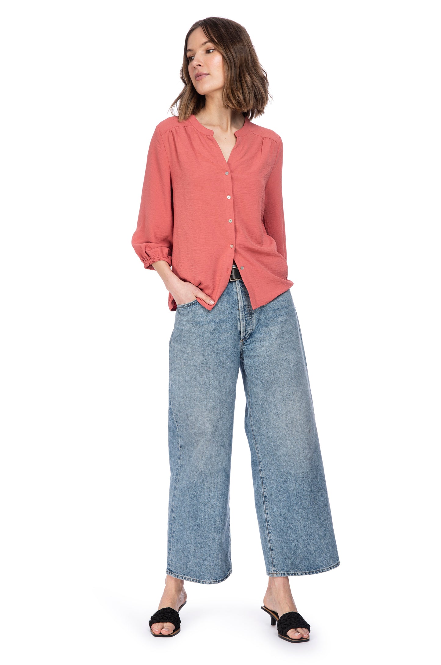 A woman stands confidently wearing a casual outfit that consists of a loose-fitting coral River Button Up Blouse made from 100% Polyester, wide-leg denim jeans, and black open-toe sandals. Brand Name: B Collection by Bobeau