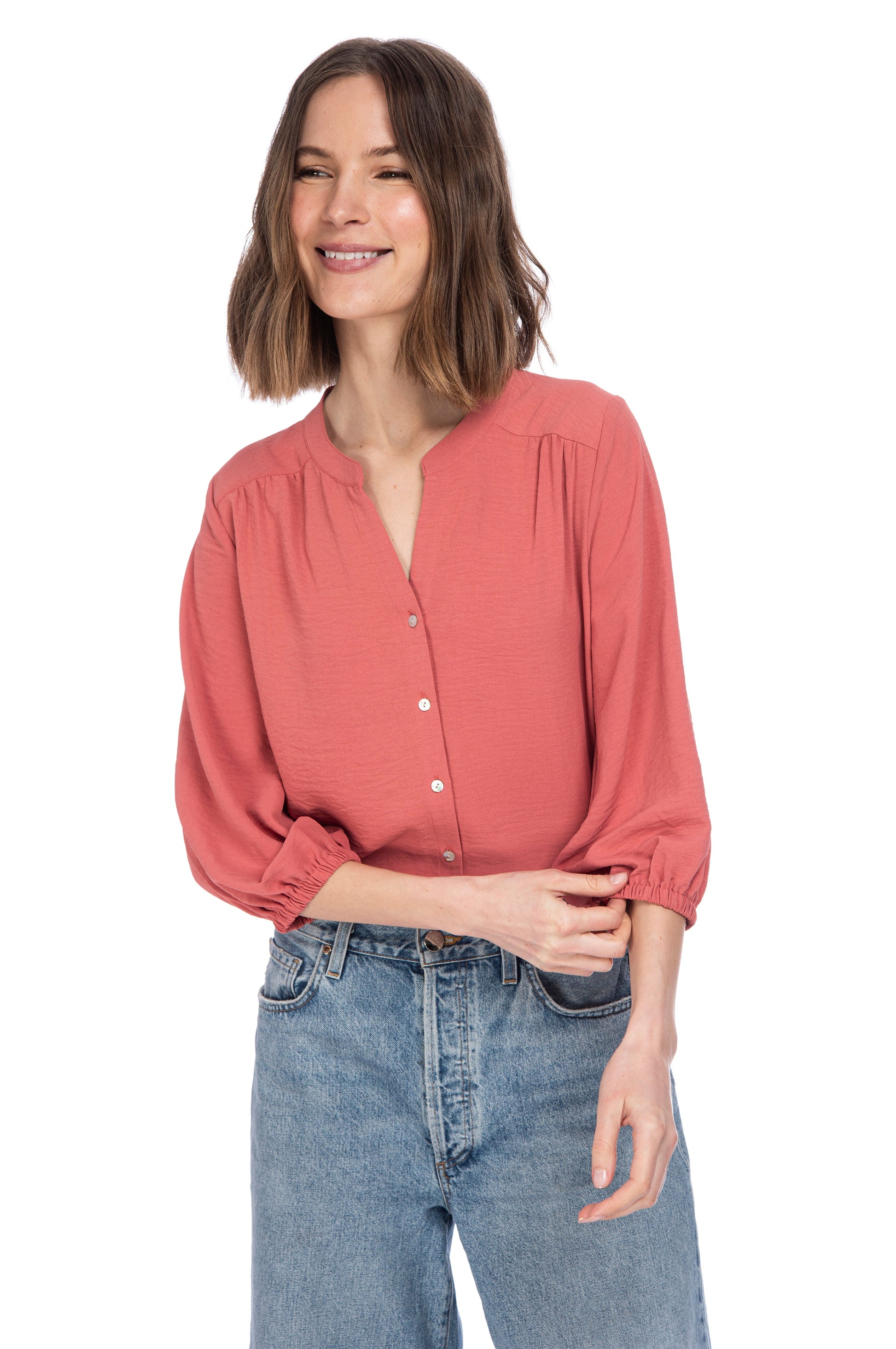 A woman with a subtle smile wearing a B Collection by Bobeau River Button Up Blouse in peach and blue jeans, standing against a white background.