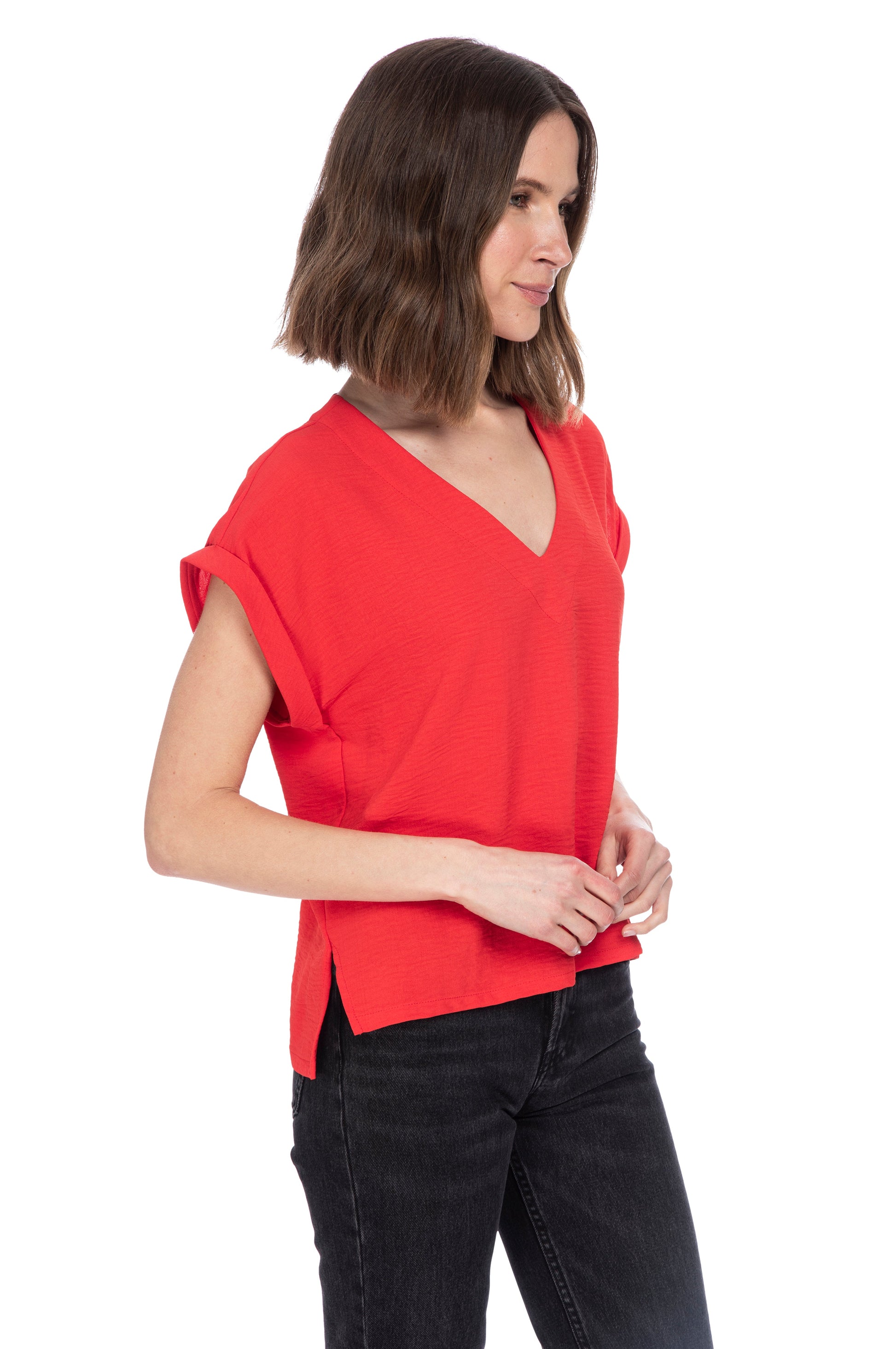 A woman in a casual red V Neck Relaxed Woven Tee made of 100% Polyester and black jeans standing against a white background, looking to her side with a thoughtful expression.