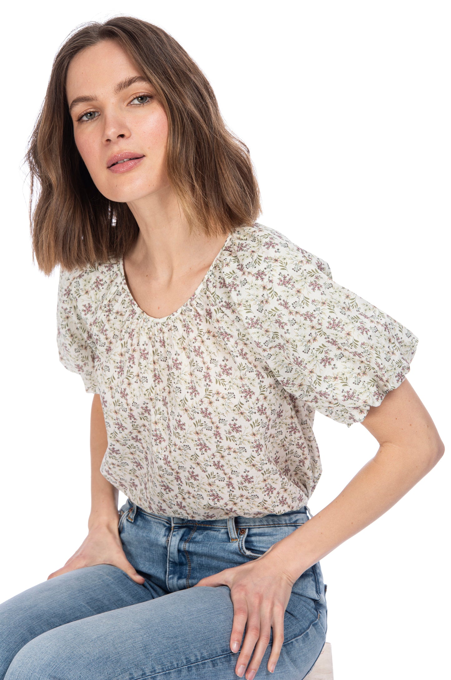 A woman with shoulder-length brown hair, wearing a crewneck B Collection by Bobeau Bubble Sleeve Cotton Top with elastic sleeves and blue jeans, casually poses while leaning forward slightly with a neutral expression on her face.