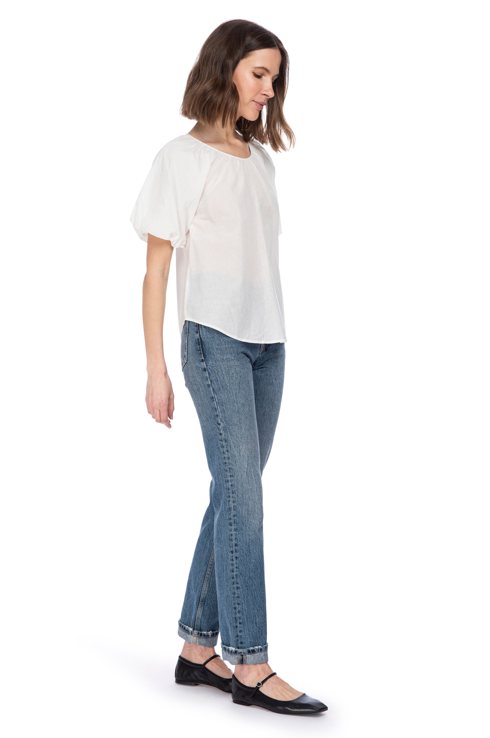 A woman in casual style wearing a white, 100% cotton B Collection by Bobeau Bubble Sleeve top, blue jeans, and black flats, posing with her head turned to one side.