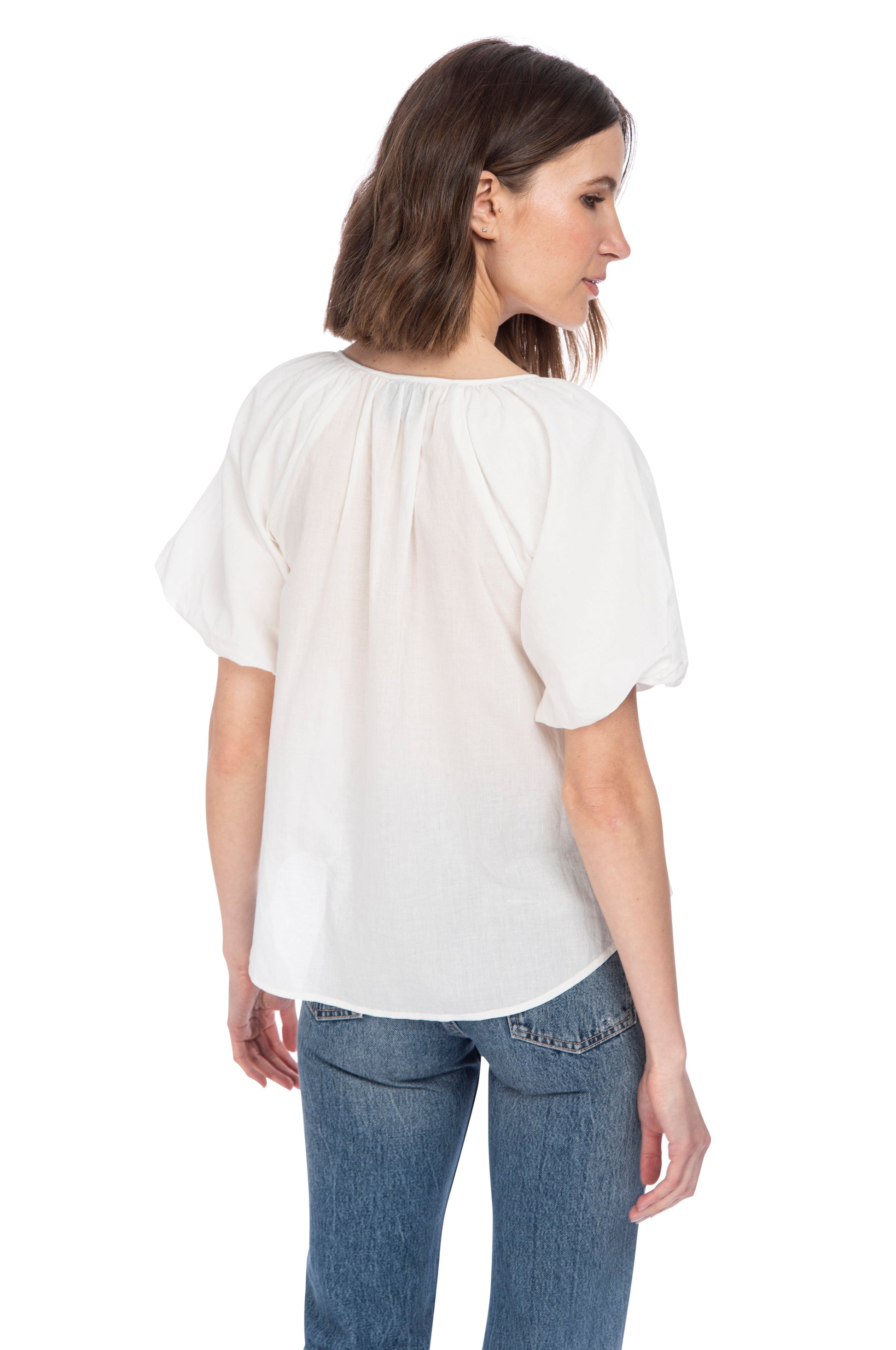 A woman seen from behind, wearing a casual cream-colored B Collection by Bobeau Bubble Sleeve Cotton Top and blue jeans, looking to the side.