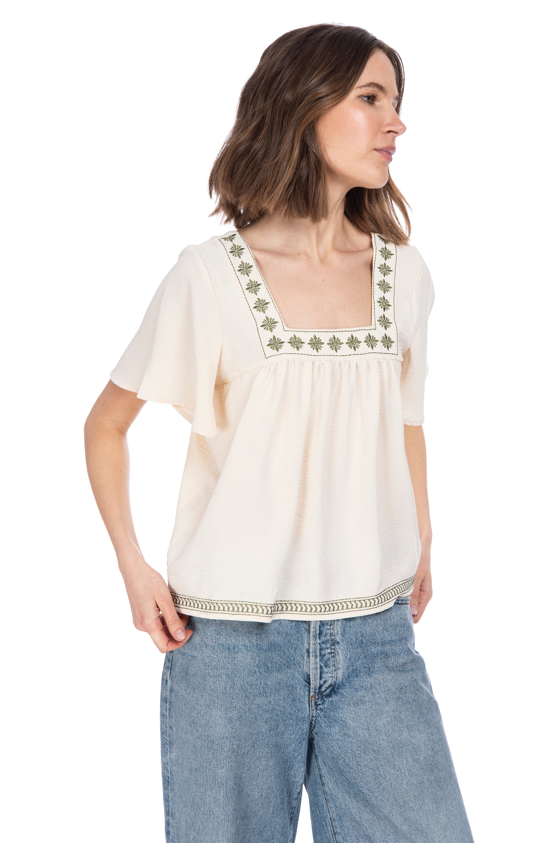 A woman in a casual off-white Square Neckline Boho Top with decorative green embroidery detail at the neckline and hem, paired with classic blue jeans, standing against a white background. (B Collection by Bobeau)