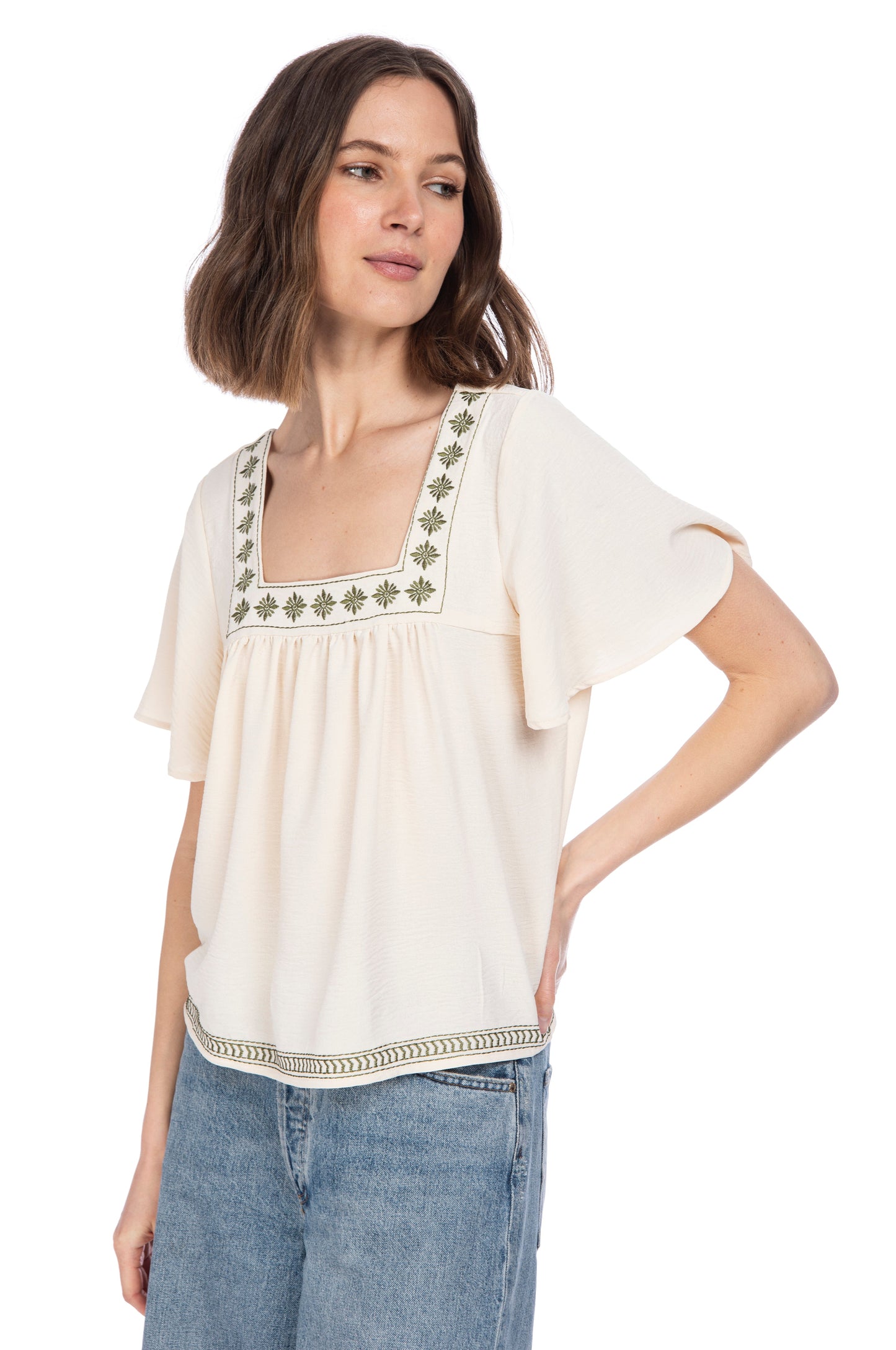 Fashionable young woman posing in a casual chic cream Square Neckline Boho Top with green embroidery detail and blue denim jeans from B Collection by Bobeau.