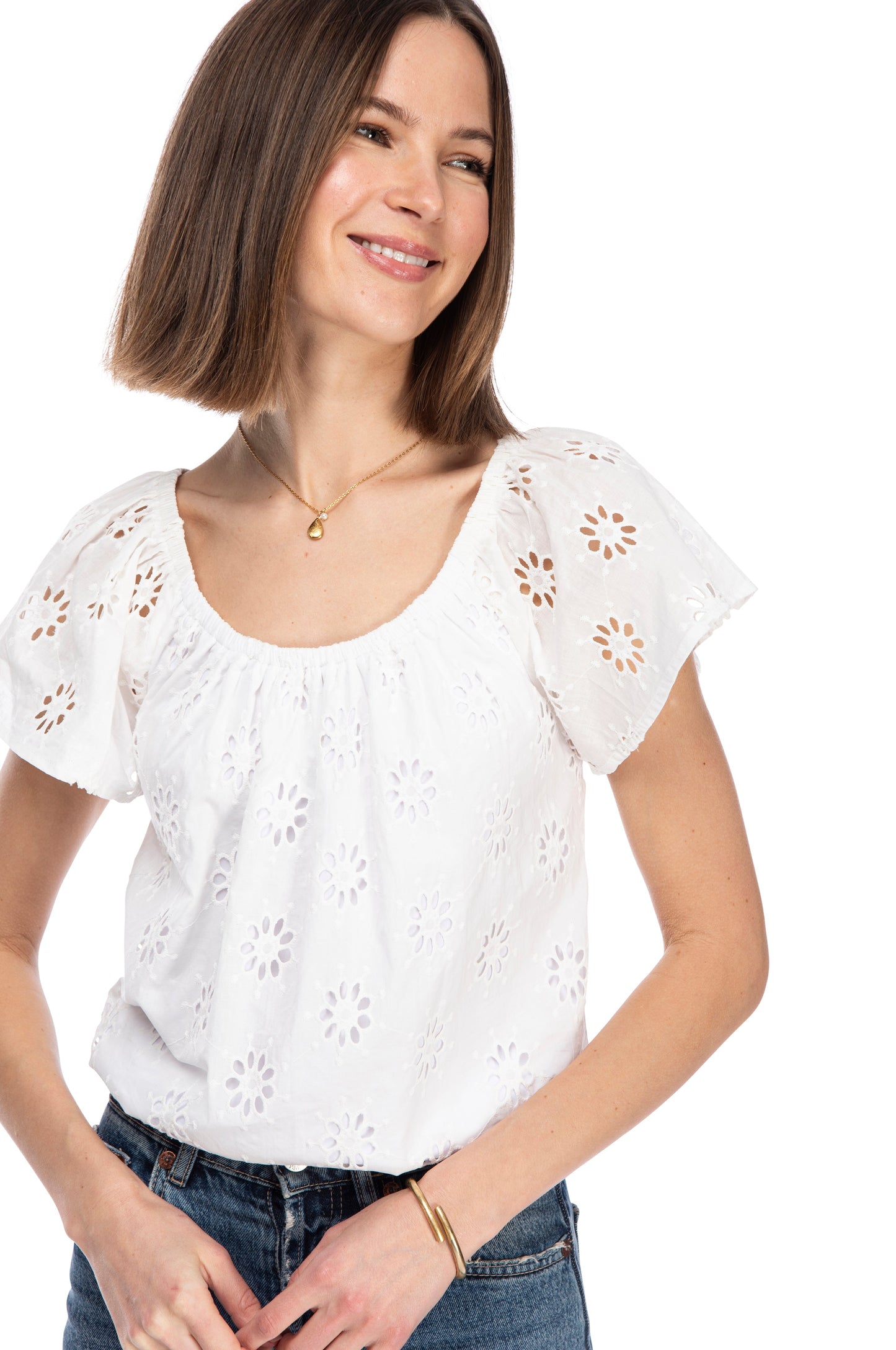A smiling woman wearing a white eyelet SS PEASANT TOP by B Collection by Bobeau with an elastic neckline and denim jeans, looking off-camera with a relaxed and friendly demeanor.