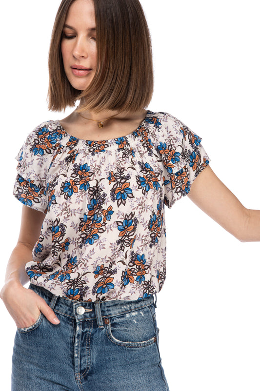 A woman in a SS TUNNEL ELASTIC TOP floral blouse by B Collection by Bobeau with an elasticated neckline and denim jeans posing with one hand on her hip.