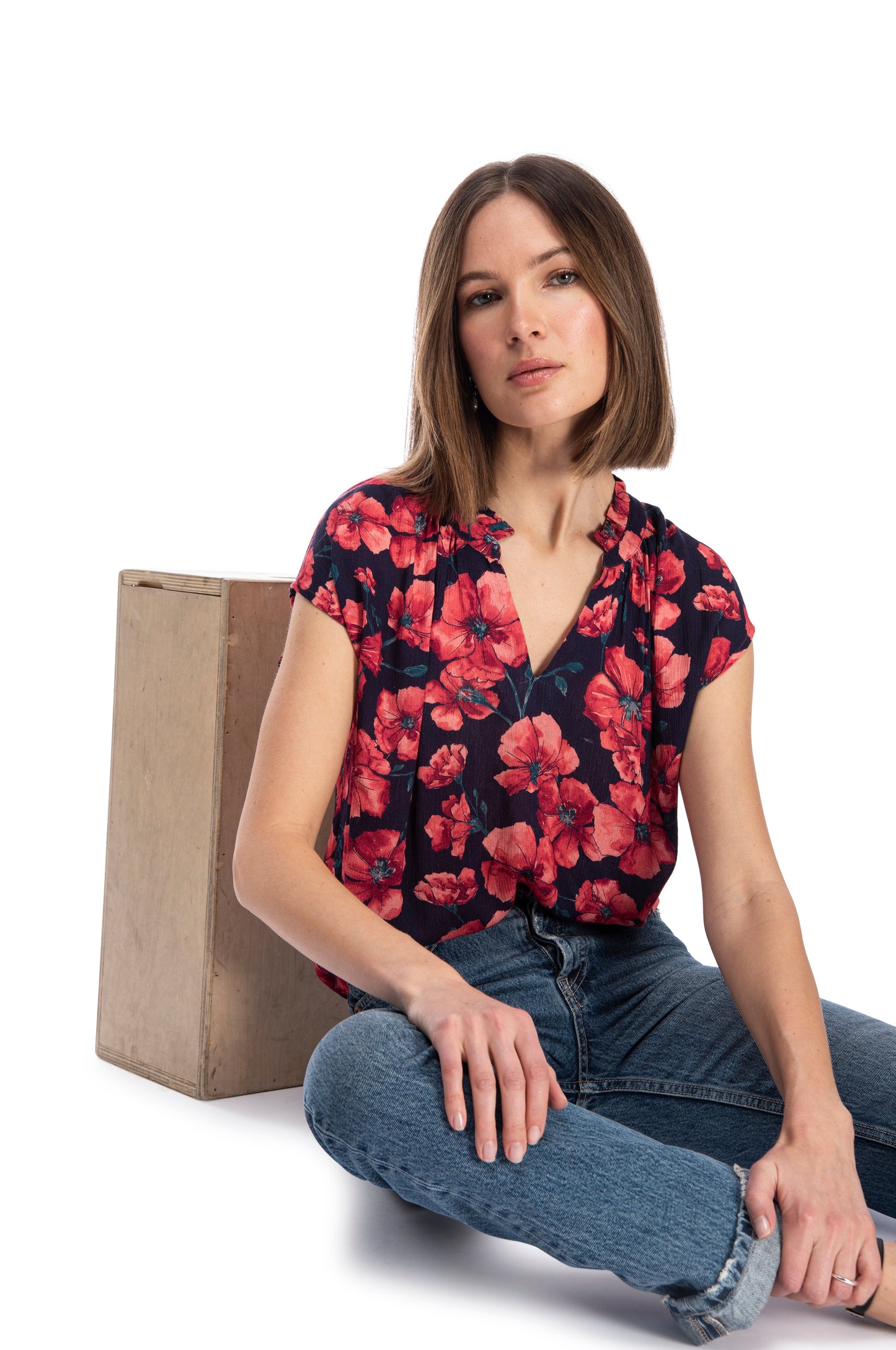 A woman in a red floral short sleeve Ruffle Neck Top from B Collection by Bobeau and blue jeans sitting casually next to a wooden box on a white background.