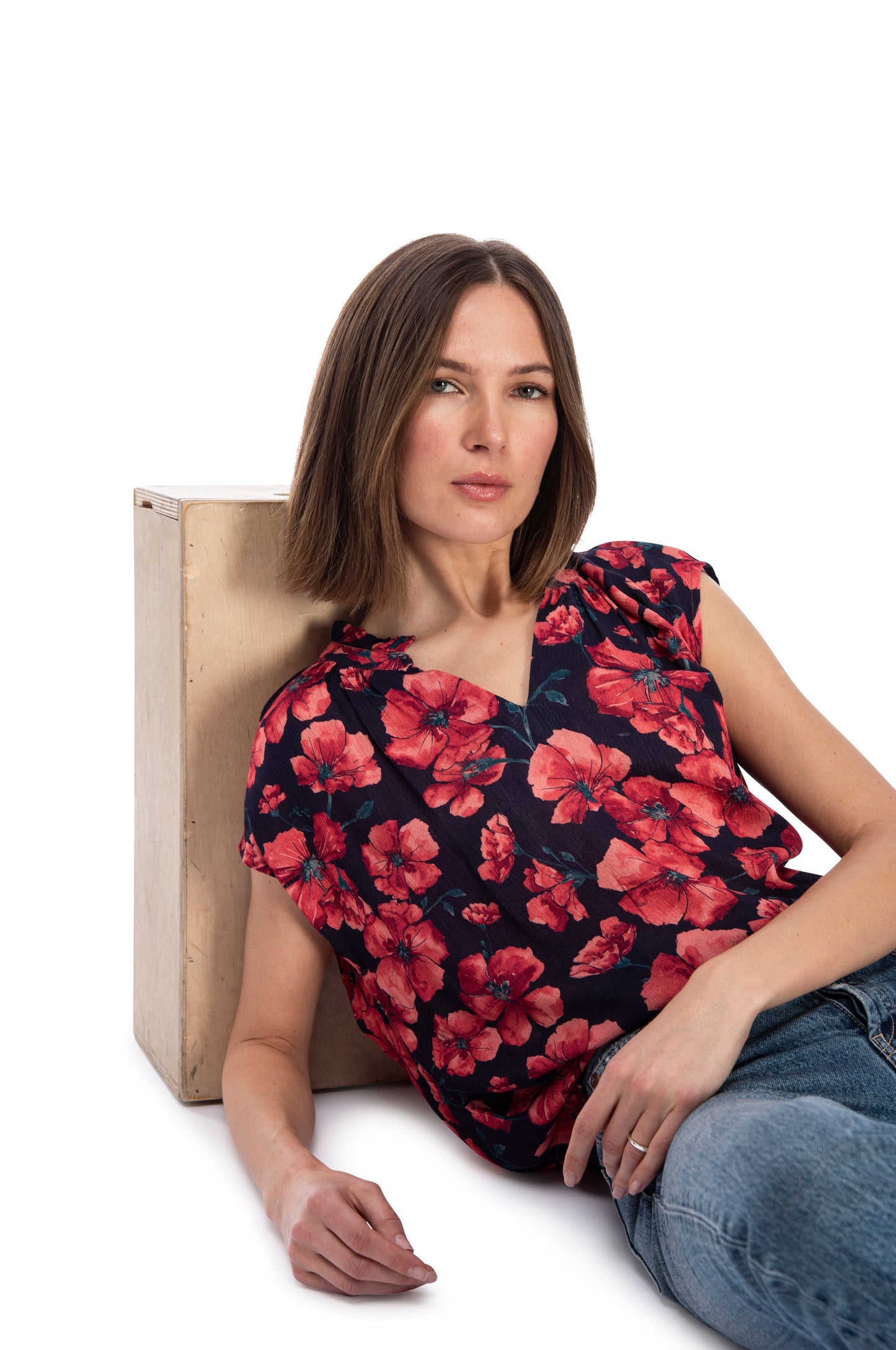 A woman in a short sleeve, floral blouse with a B Collection by Bobeau Ruffle Neck Top and jeans sitting casually next to a wooden box against a white background.