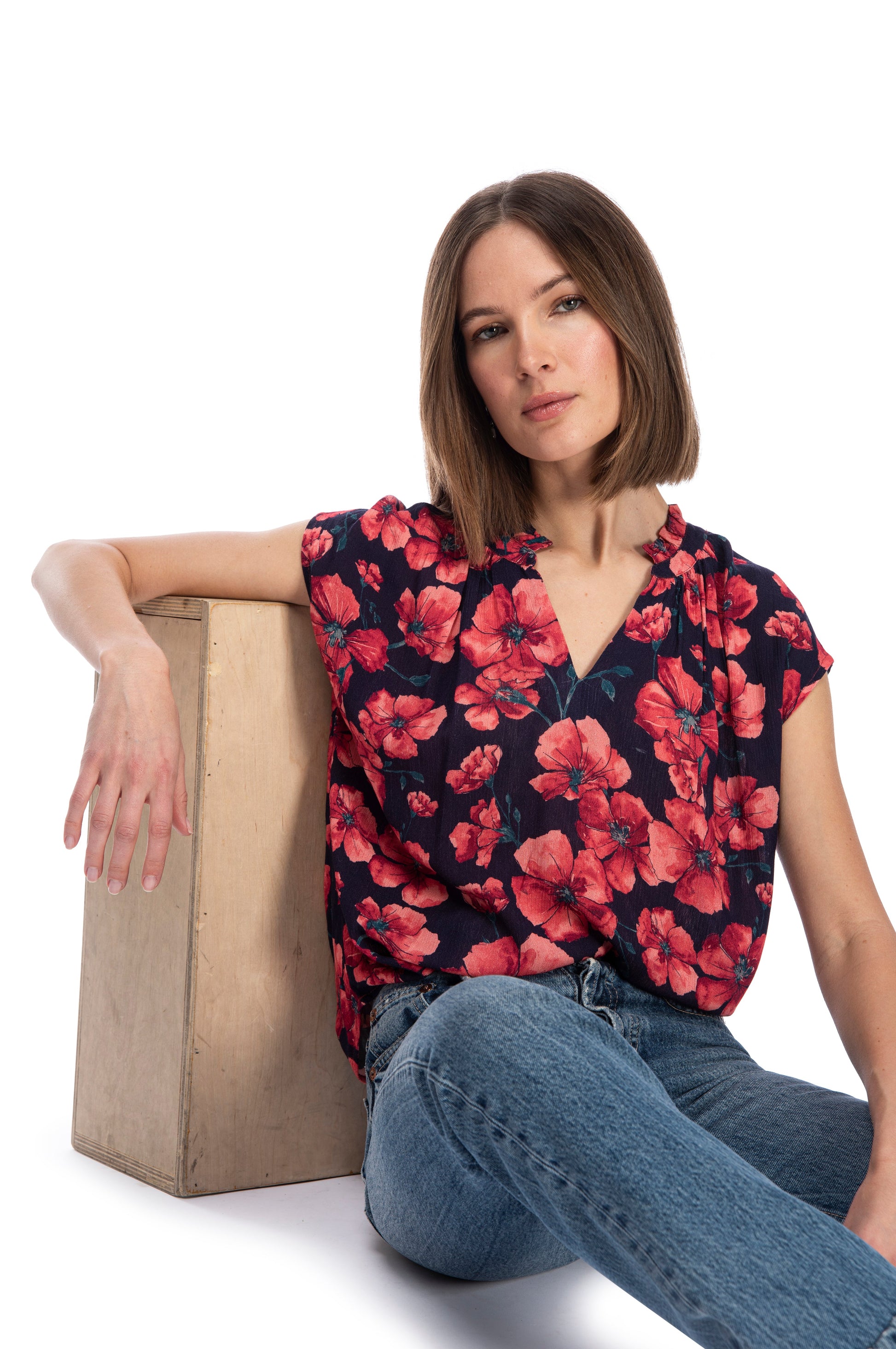 A woman in a short sleeve, floral RUFFLE NECK TOP from B Collection by Bobeau and jeans sitting casually next to a wooden box, with a relaxed and confident posture, against a white background.